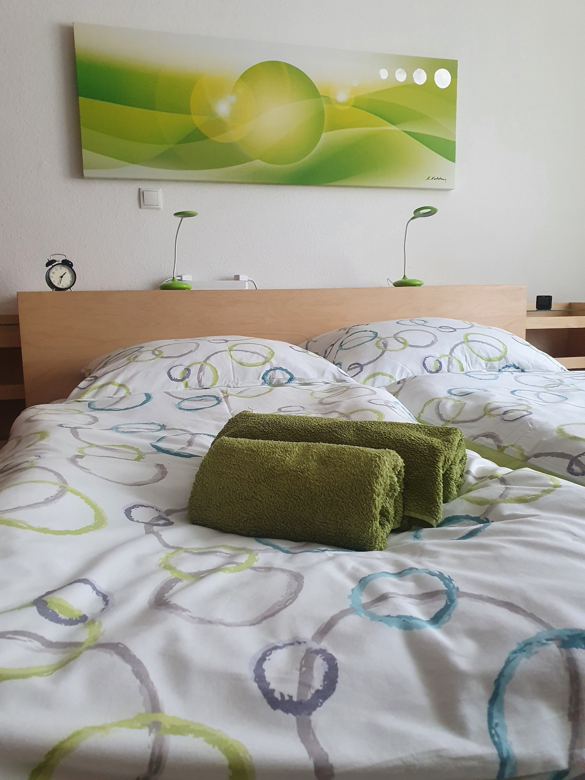 A sleeping area features a wooden bed with colorful bedding adorned with circular patterns. Two bedside lamps in green provide soft illumination, while a vibrant artwork hangs above. Fresh green towels are neatly placed at the foot of the bed, enhancing the welcoming ambiance.