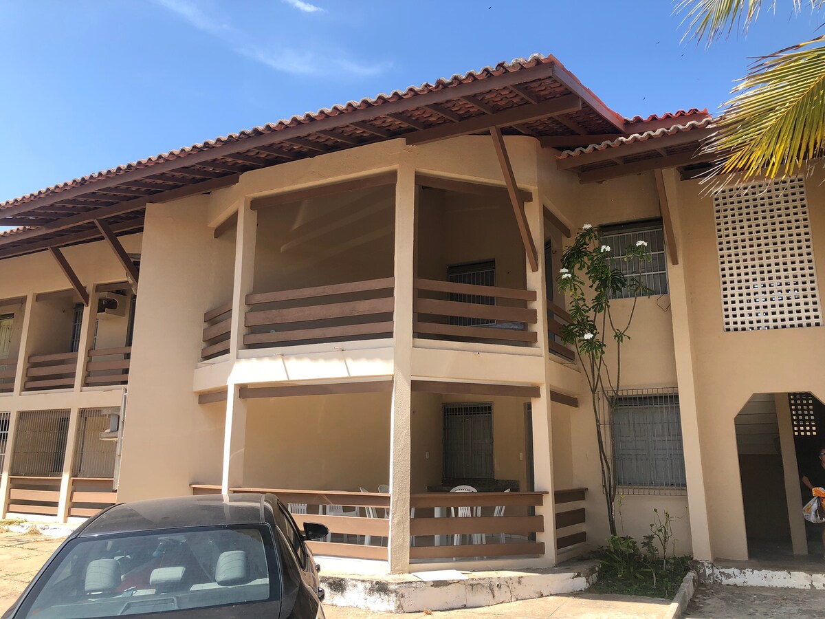 The exterior of a two-story apartment building is presented, featuring balconies with wooden railings. A parked car is visible in the foreground, and a palm tree is seen on the right side. Sunlight brightens the scene under a clear blue sky.