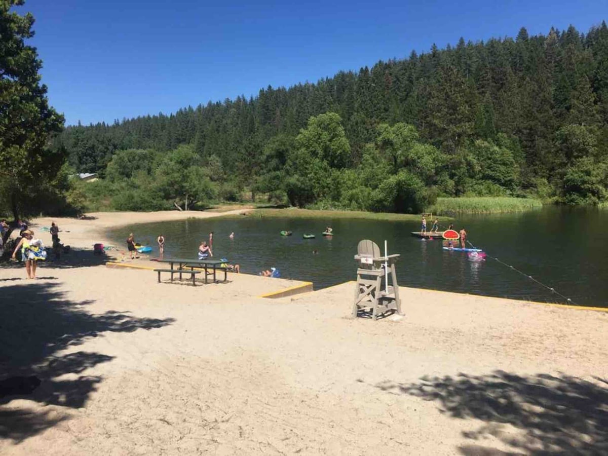 A sandy beach area is depicted, featuring a calm lake surrounded by lush greenery and trees. Several people can be seen enjoying the water on inflatable floats, while a picnic table sits on the shore. A lifeguard chair is positioned nearby, overlooking the swimming area.