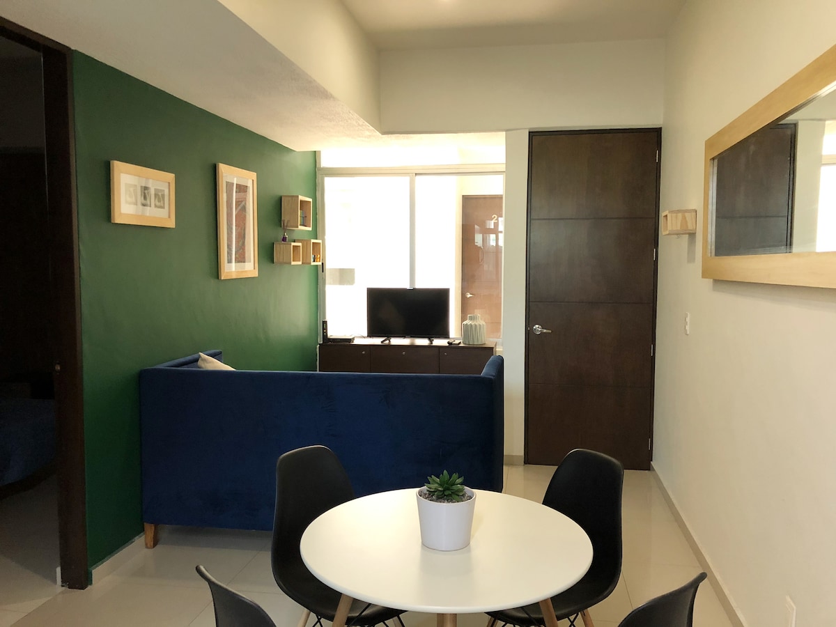 A modern living space featuring a deep blue sofa and a white round dining table surrounded by black chairs. A television stands on a light wooden console, with natural light streaming in through large windows. Green accent walls add depth to the room.