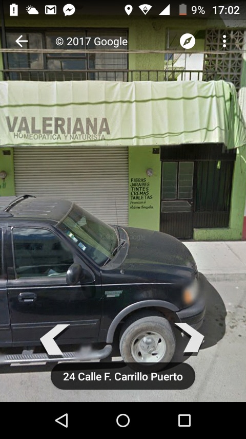 A black pickup truck is parked on the street in front of a green building featuring a sign that reads 'VALERIANANA HOMEOPATICA Y NATURISTA.' A partially closed metal roller door is visible alongside a wall adorned with writing.
