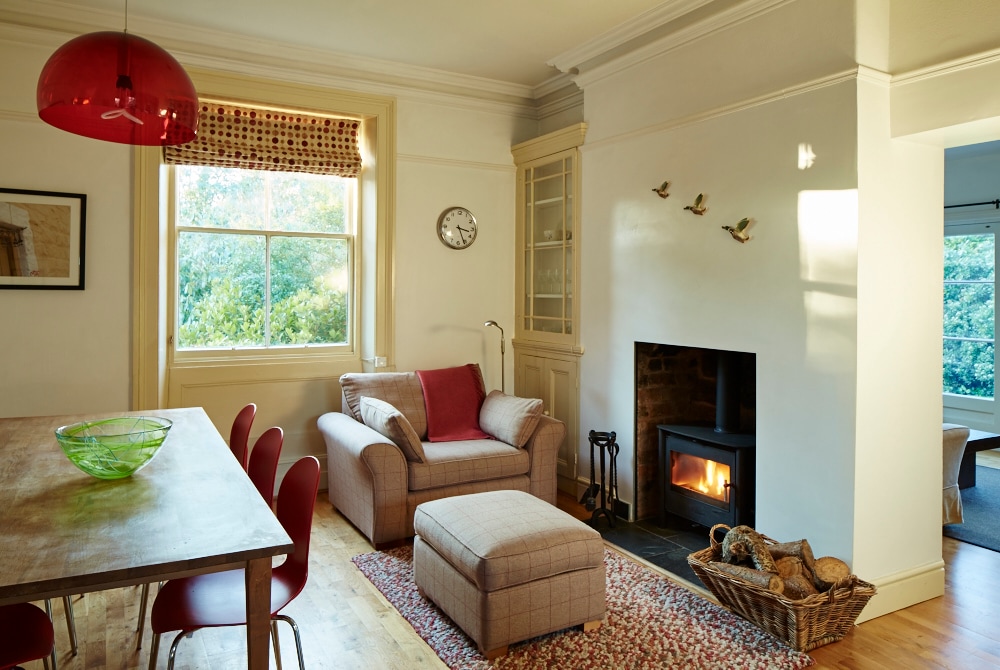 A comfortable living area is highlighted by a cozy armchair beside a warm fireplace. A dining table with red chairs is positioned nearby, complemented by a decorative rug. Natural light enters through a large window adorned with a shade, enhancing the overall inviting ambiance.