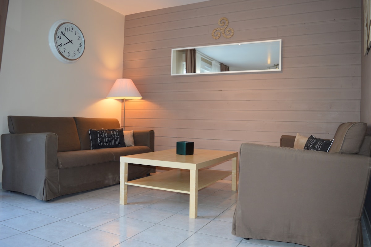A living area is featured with two brown sofas positioned around a light wood coffee table. A floor lamp provides soft lighting, and a round wall clock hangs prominently. A mirror reflects the space, enhancing the overall brightness.