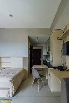 A cozy sleeping area features a spacious bed covered with a light-colored bedding set. An adjacent workspace is equipped with a minimalistic desk and a chair. The room includes a kitchenette with contemporary cabinets and a television mounted on the wall.