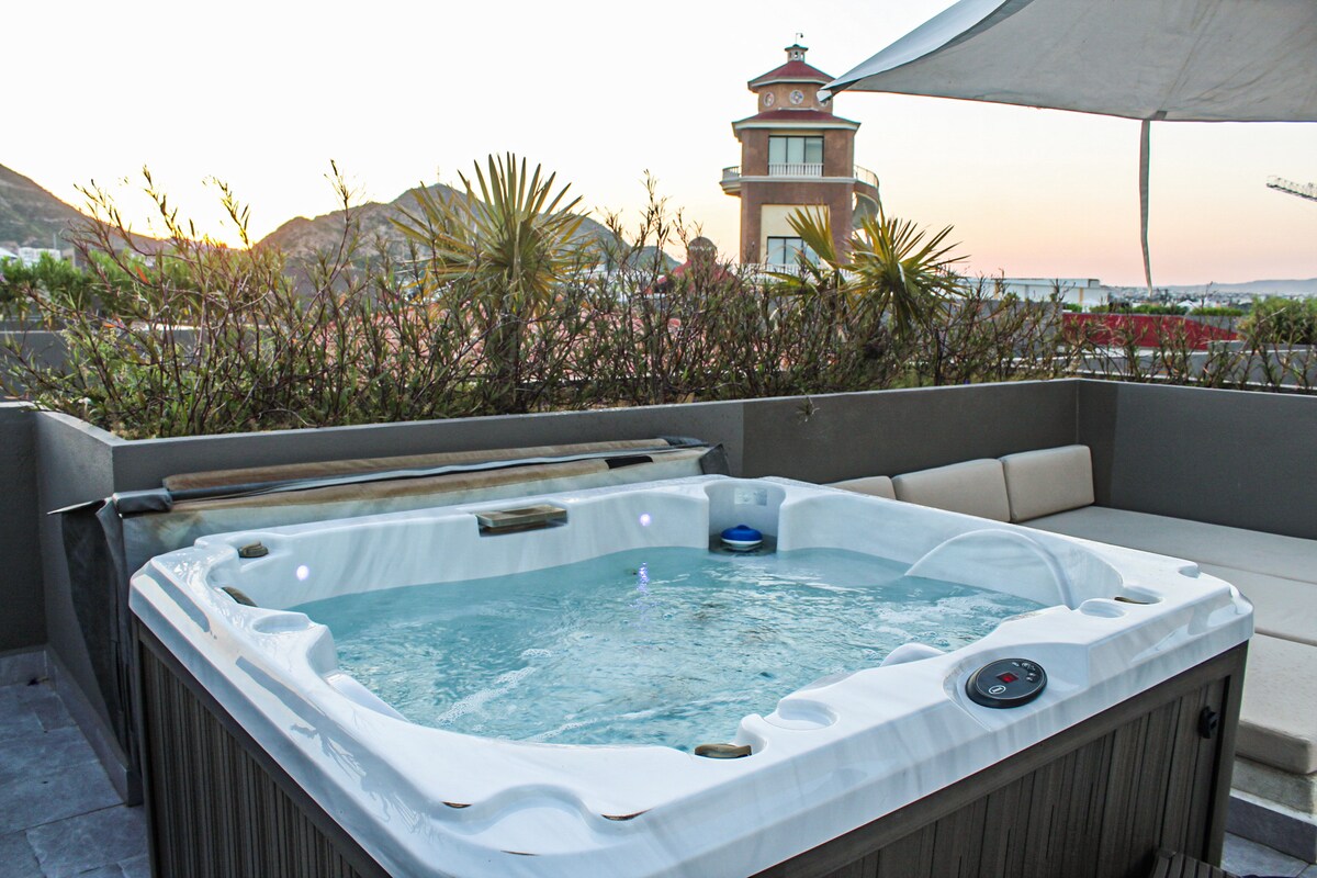 A hot tub with bubbling water is situated on a private terrace, surrounded by greenery. Soft seating is positioned nearby, providing a relaxing spot to enjoy the sunset views over the hills and surrounding architecture.