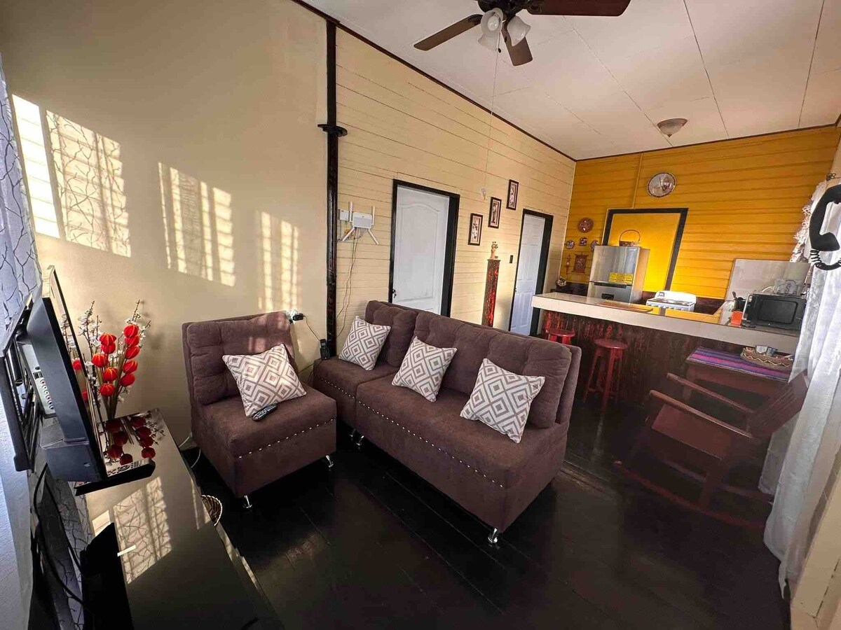 A cozy living area features a pair of brown sofas with decorative cushions, accompanied by a television and a small coffee table. Natural light pours in through the large window, illuminating the space. A dining area with bar stools is visible in the background.
