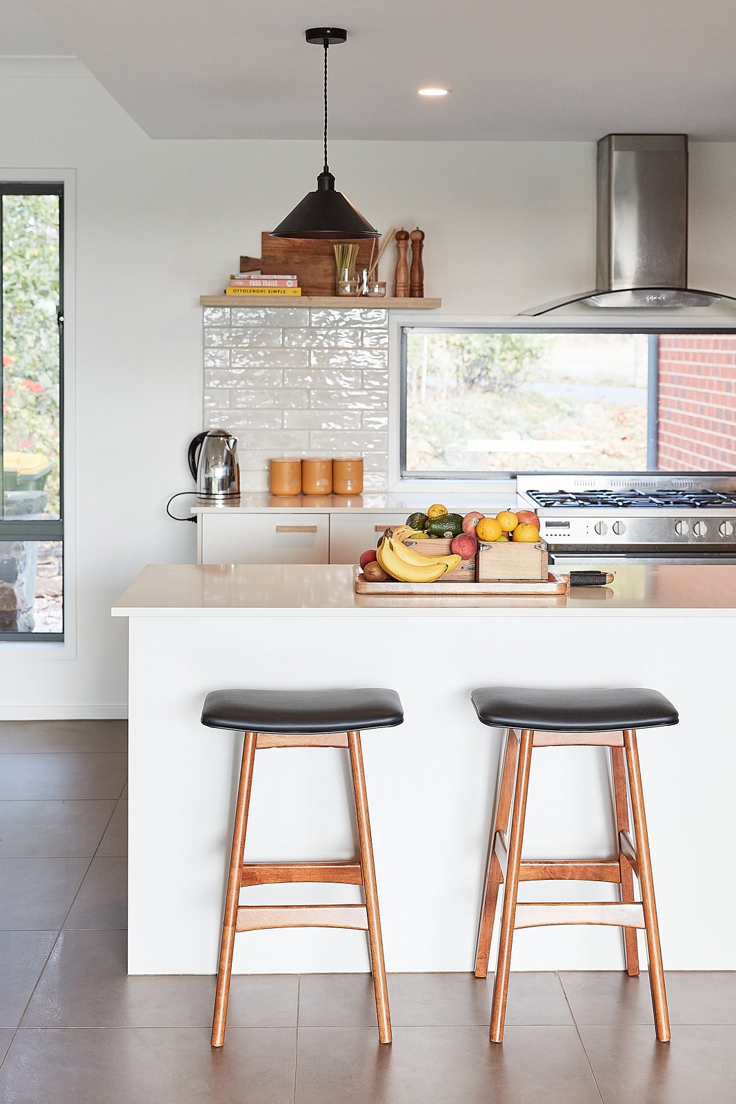 A modern kitchen features a central white island with bar stools positioned at one side. Fruits are arranged on a wooden tray, while kitchen appliances and utensils are displayed on shelves, with natural light filtering through the window.