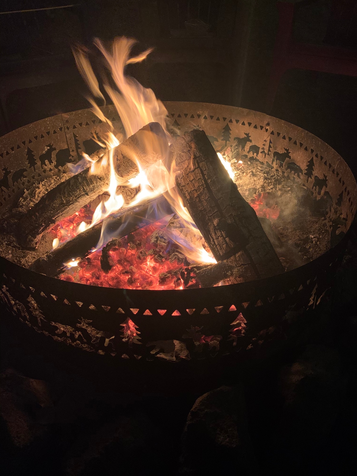 A round fire pit, intricately designed with cut-out patterns, contains a lively fire. Logs are burning brightly, casting warm light and shadows, while glowing embers are visible amidst the ashes, creating an inviting focal point for gatherings.