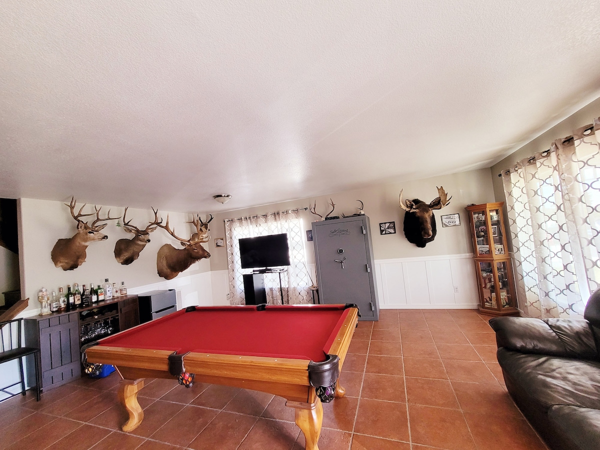 A spacious game room features a red felt pool table as the central focus. The walls are adorned with mounted antlers, while a flat-screen television is mounted above a wooden bar area. Natural light filters through sheer curtains, and a large safe stands against the wall.