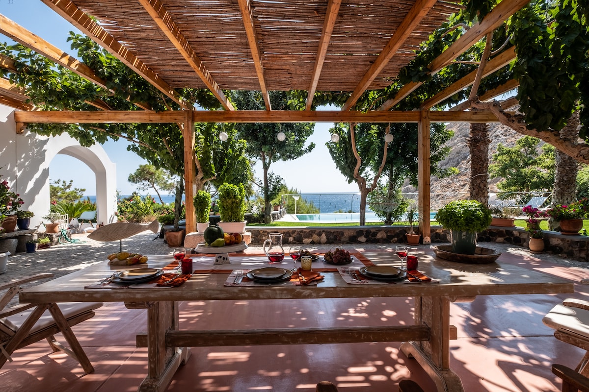 A shaded outdoor dining area features a long wooden table set with plates and drinks, surrounded by lush greenery. The view of the sea is visible in the background, enhancing the natural setting. Soft light filters through the trellis above, creating a serene ambiance.