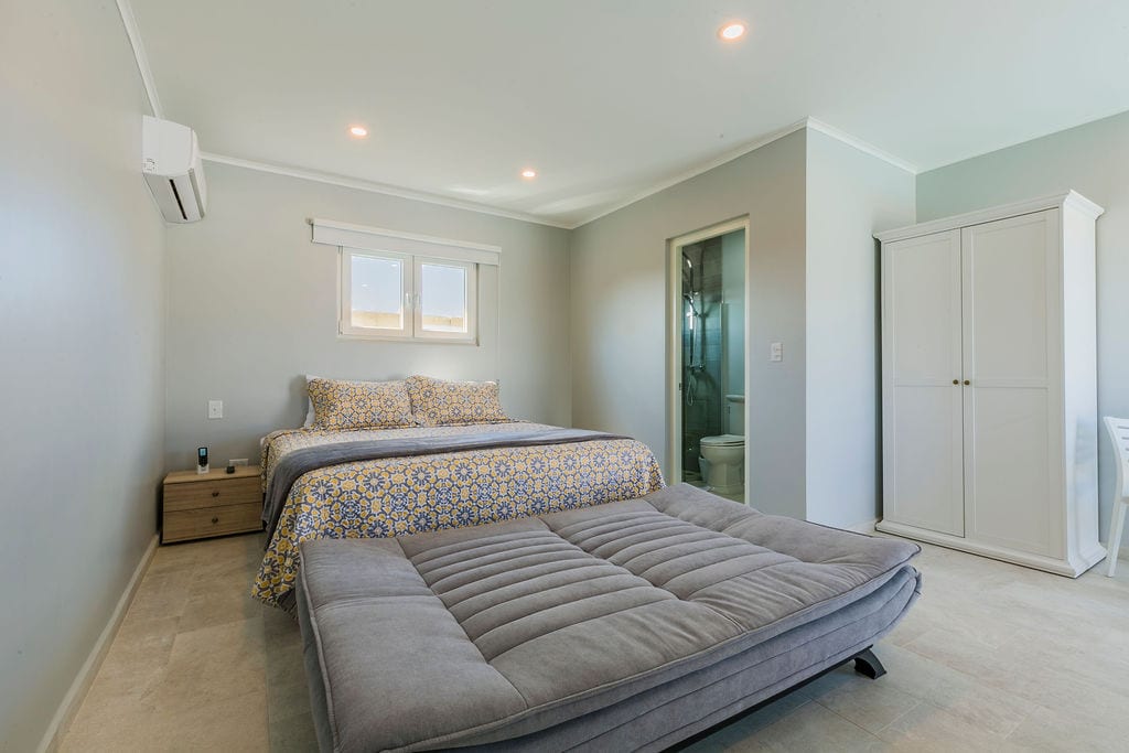 A spacious bedroom is displayed, featuring a queen bed with a patterned bedspread and an additional fold-out mattress on the floor. Natural light enters through a window beside the bed, illuminating the light grey walls and tiled flooring. A closet stands in the corner, with a bathroom accessible in the background.