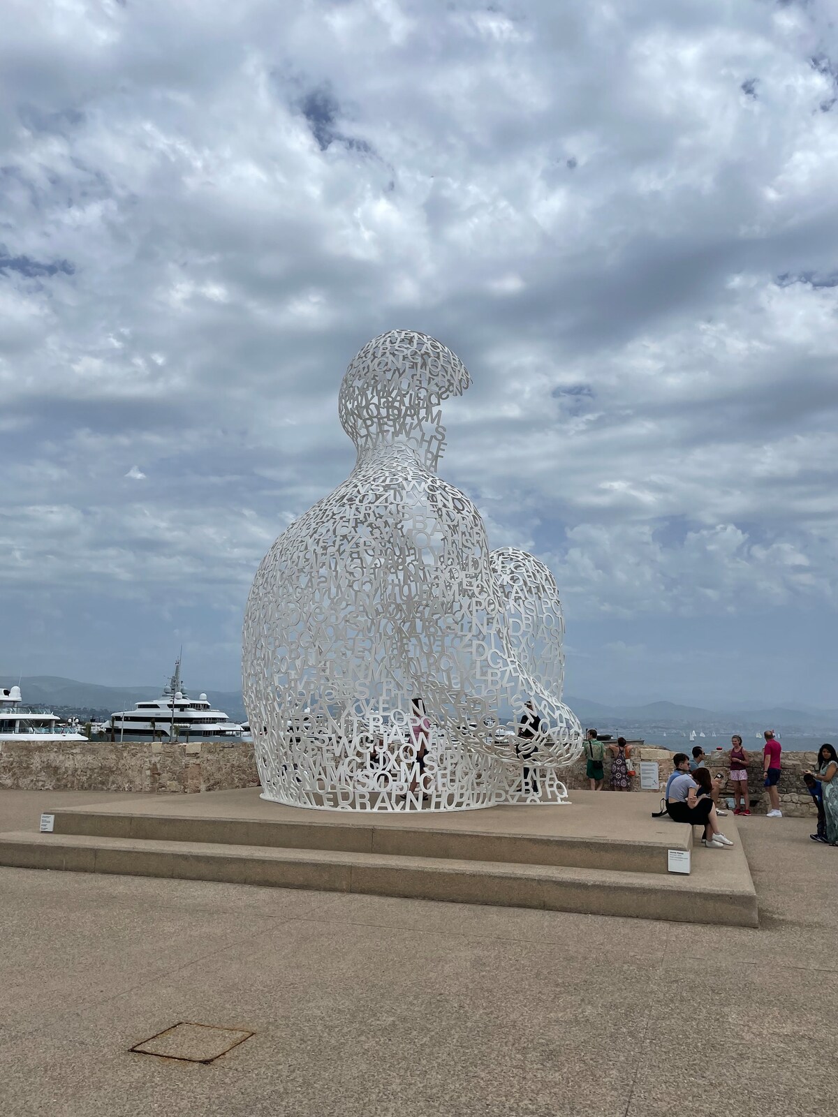 A large, intricate white sculpture is prominently displayed against a cloudy sky. Visitors can be seen admiring the piece, with views of distant yachts and the horizon creating a tranquil backdrop. The sculpture's unique design reflects a collection of letters and forms.