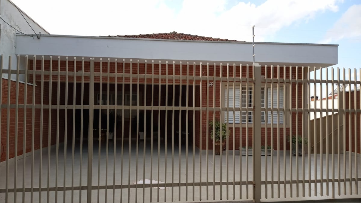 A large, fenced entrance area is revealed, showcasing a spacious driveway and classic brick facade of the house. A set of windows is visible above the fence, providing a glimpse of the interior beyond. Potted plants add a touch of greenery.