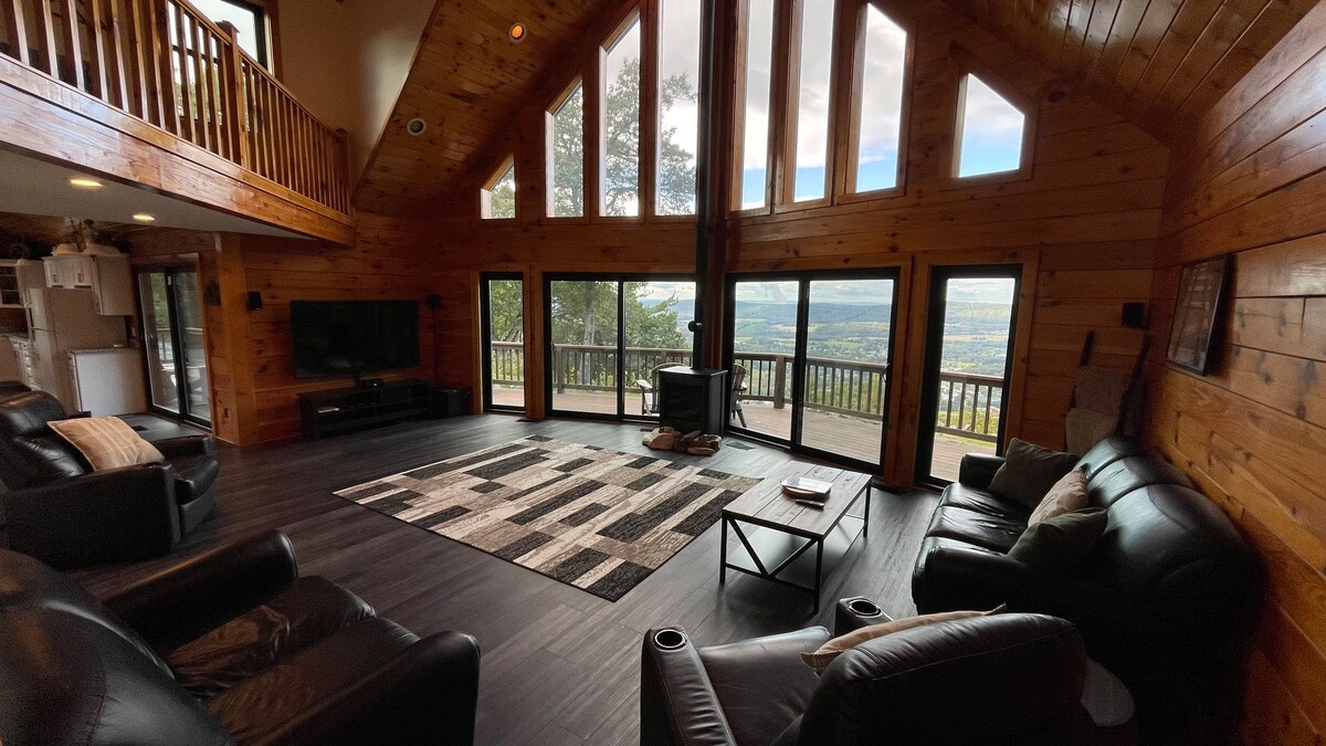 A spacious living area is surrounded by large windows offering expansive views of the valley. Dark leather seating is arranged around a geometric area rug, with a modern coffee table in the center. A wood-burning stove is positioned near the windows, creating a cozy focal point.