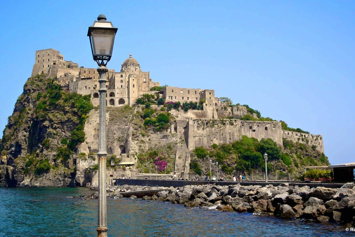 A coastal scene is depicted, showcasing a historical castle perched on a rocky hill. The structure features various levels and retaining walls, surrounded by greenery. A streetlamp stands in the foreground, contributing to the waterfront ambiance.