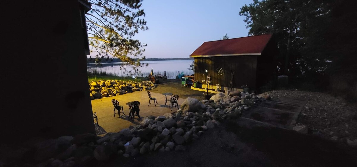 An outdoor patio area is illuminated softly in the evening light, featuring several metal chairs arranged near a stone border. The calm lake is visible in the background, surrounded by trees, indicating a serene atmosphere.