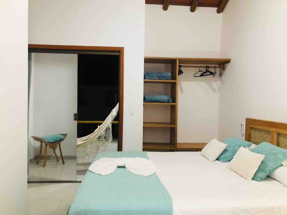 The sleeping area features a spacious bed with light blue and white accents. A hammock is visible in the background, along with a small wooden chair and an open shelf with folded towels. Natural light enters through the open door.