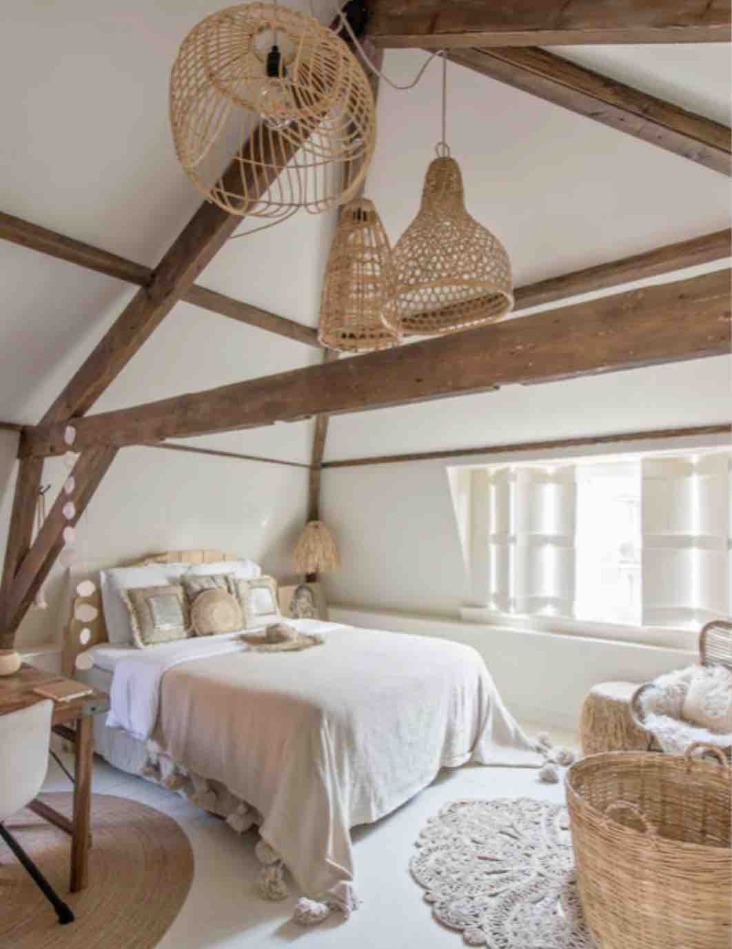 A bright bedroom features a spacious bed covered with a light-colored blanket and decorative pillows. Woven pendant lights hang from the ceiling beams. Natural light enters through a window adorned with wooden shutters, illuminating the cozy seating area with woven baskets.