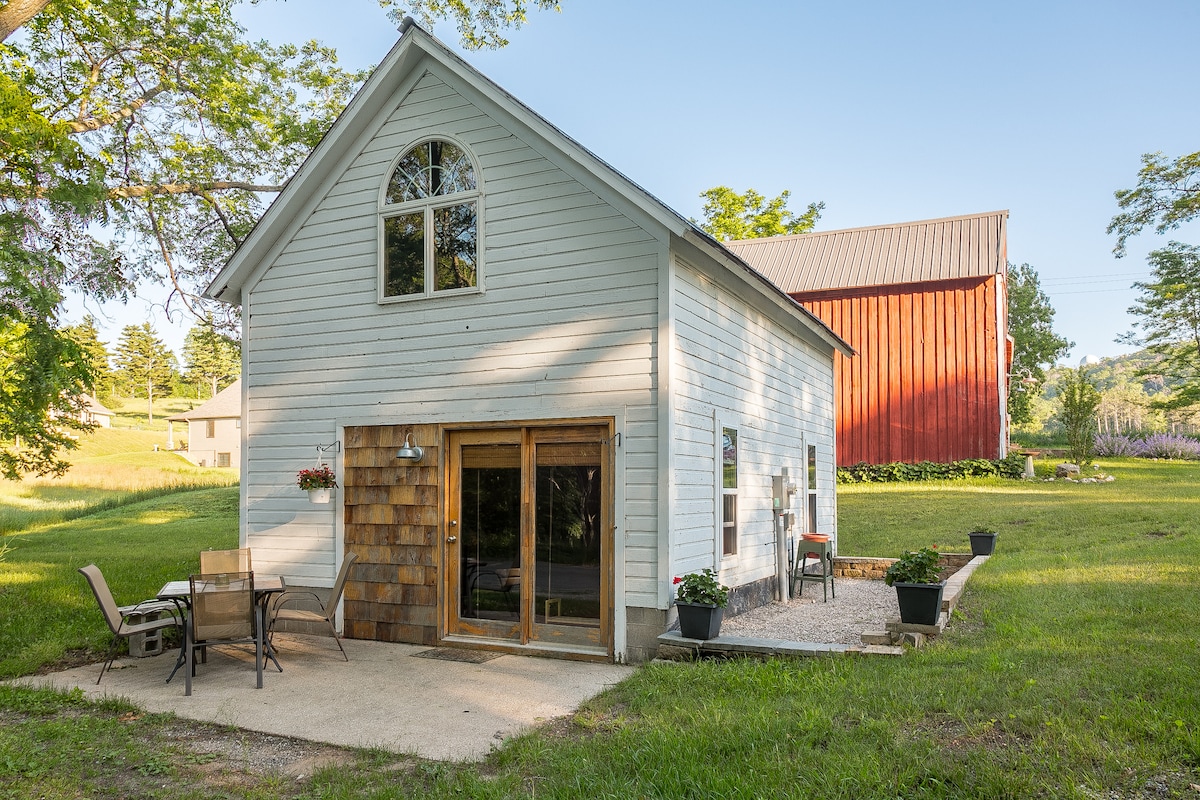 The exterior of the Carriage House is characterized by a light-colored facade and large glass doors, allowing natural light to fill the space. Outdoor seating is arranged on a stone patio, surrounded by greenery, with a red barn standing in the background.