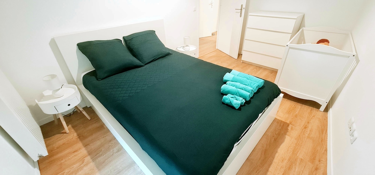 A simple bedroom features a spacious bed with dark bedding, complemented by two untucked green towels neatly arranged on the side. A sleek white nightstand is positioned alongside the bed, and a dresser stands against the wall. A baby crib is discreetly placed nearby.