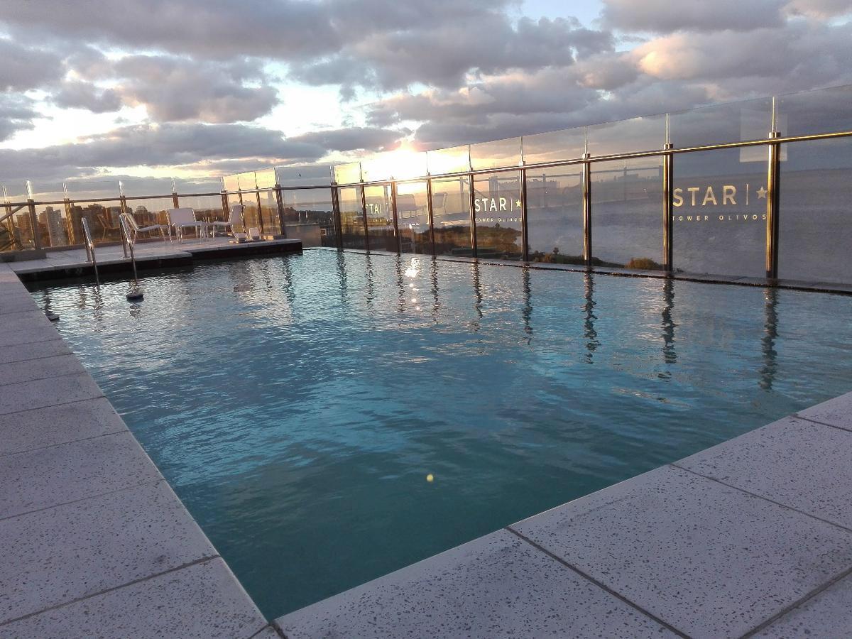 An outdoor swimming pool reflects the soft hues of the sky at sunset, framed by a transparent glass barrier. Surrounding areas are paved, with a few lounge chairs positioned for comfort and relaxation. The horizon features a tranquil view of the water.