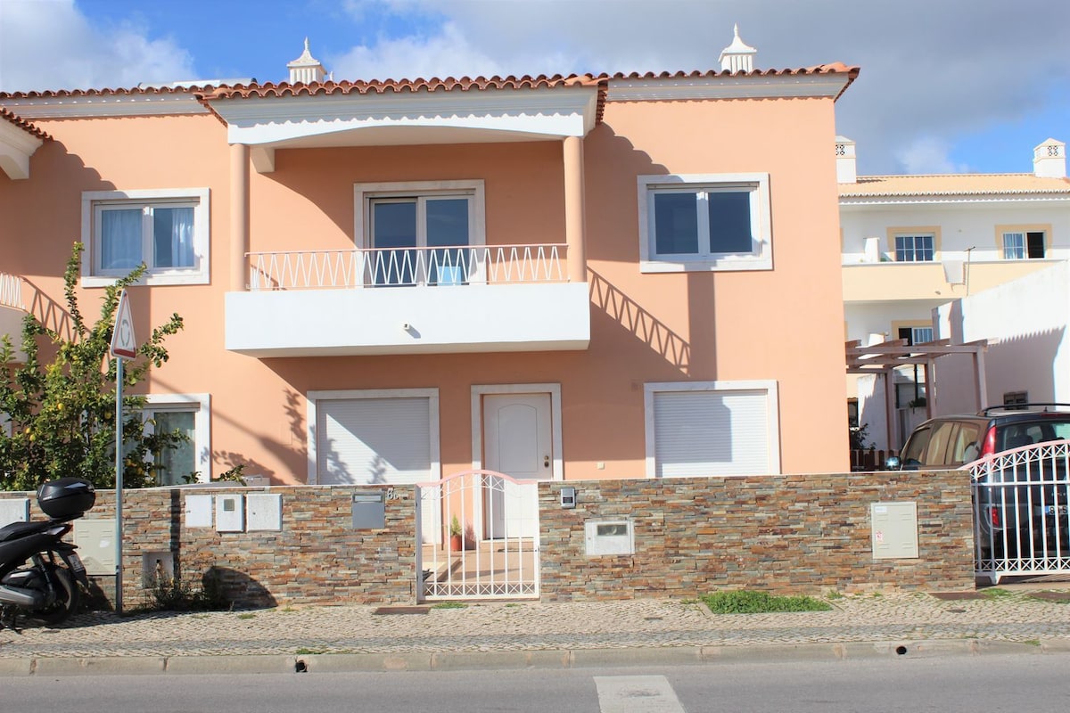 A two-story villa features a warm pink exterior, framed by white window shutters. A decorative stone wall surrounds the property, with a gated entrance leading to the front door. The sidewalk is lined with small plants, and parked vehicles can be seen nearby.