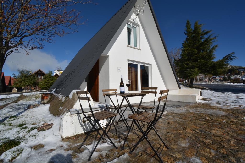 A modern triangular-shaped house is seen against a clear blue sky, with snow covering the ground. A small outdoor seating area is furnished with a wooden table and four folding chairs, complemented by a bottle of wine positioned on the table.