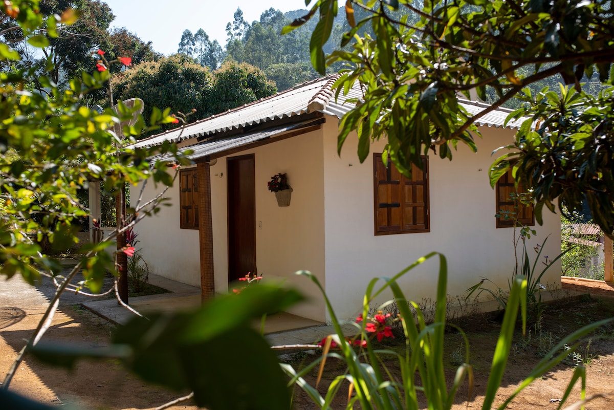 A single-story white house is framed by lush green foliage and colorful flowers. Wooden windows are visible, adding a natural touch to the exterior. The surrounding landscape features trees and hills in the background, enhancing the tranquil setting.