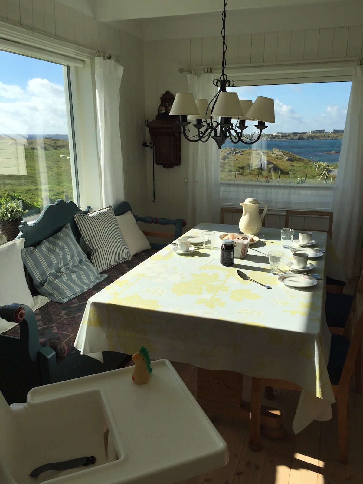 A dining area features a large table set with dishes and glassware. Light-colored curtains frame the windows, providing views of the surrounding landscape. A chandelier hangs overhead, and a high chair is positioned nearby, creating a cozy space for shared meals.