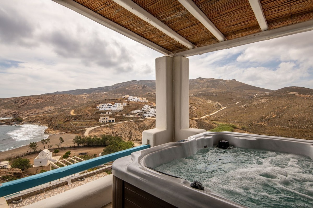 A private jacuzzi is positioned on the terrace, offering scenic views of the surrounding hills and coastline. The structure's overhang provides shade, while the bubbles of the jacuzzi create a tranquil atmosphere. Soft, cloudy skies are visible, enhancing the serene setting.