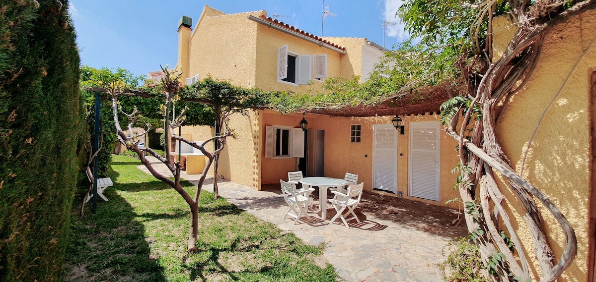 A charming outdoor area is enclosed by yellow walls, featuring a stone patio with a round table surrounded by four white chairs. Lush greenery, including trellises with vines, frames the space, creating a tranquil ambiance suitable for relaxation.