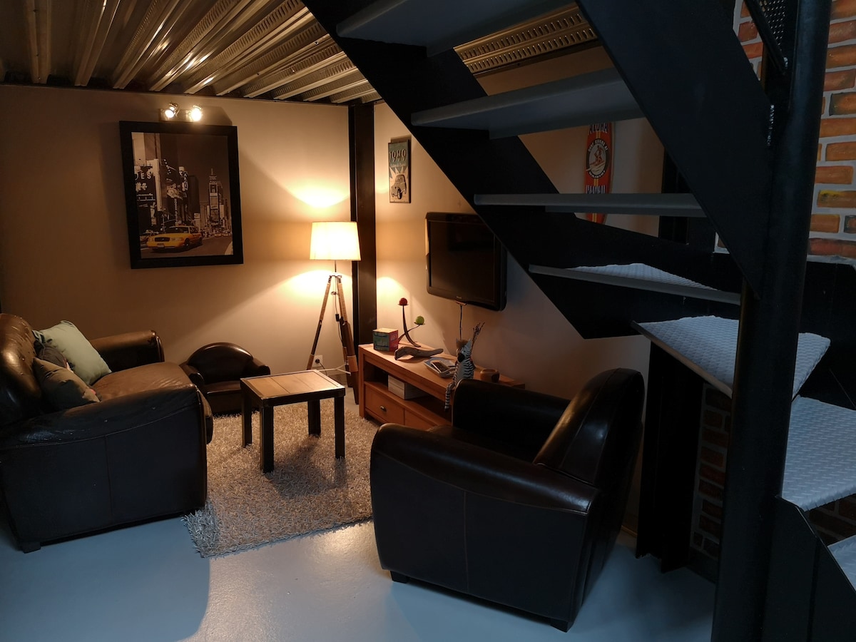 A cozy living area features comfortable leather seating, a wooden coffee table, and a soft rug. A floor lamp provides warm light, while a wall-mounted television adds to the entertainment options. Stairs lead to the upper level, enhancing the space's functionality.