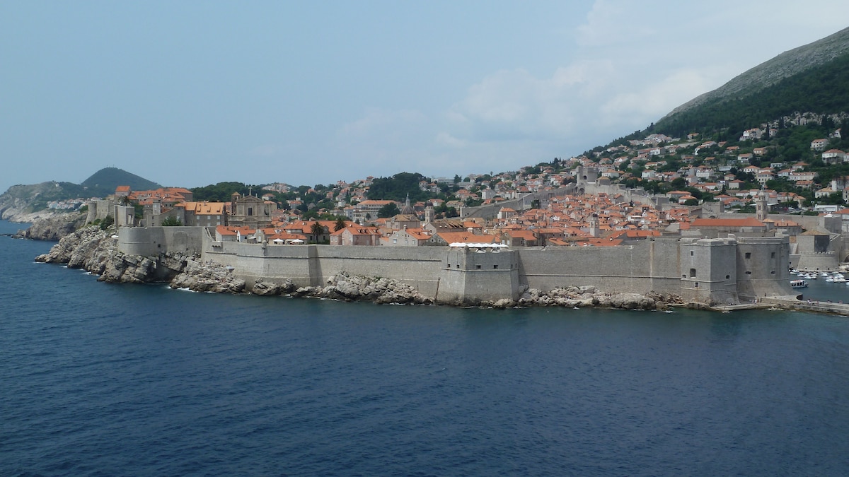 A coastal view showcases a fortified stone structure along the shore, surrounded by calm blue waters. The ancient city walls rise above the rocky coastline, while terracotta-roofed buildings and green hills are seen in the background, creating a harmonious landscape.