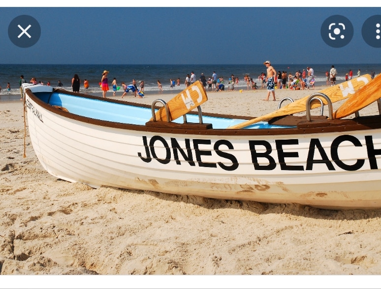 A white lifeguard boat sits on the sandy beach, with the words 'JONES BEACH' displayed prominently on its side. In the background, groups of people can be seen enjoying the beach and the ocean under a clear sky.