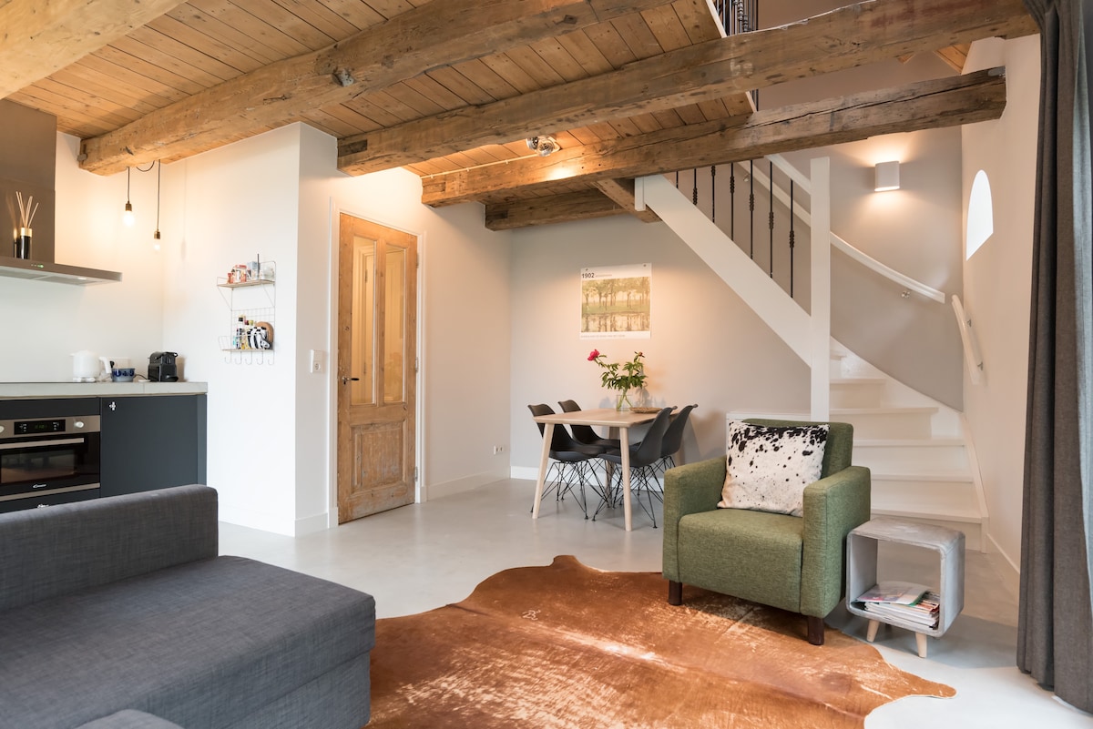 A modern living area features exposed wooden beams above, complemented by a neutral color palette. A spacious sofa faces a sleek kitchen, while a dining table with chairs is positioned near the staircase. Natural light streams in through an adjacent window.