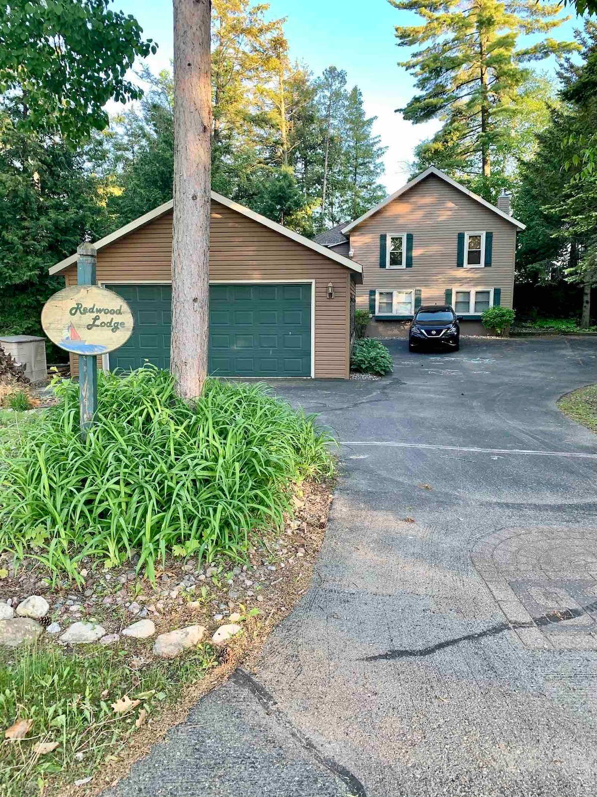 The exterior of the Redwood Lodge is displayed, featuring a two-story home with a brown exterior, green garage door, and a welcoming sign. Lush green landscaping surrounds the driveway, leading to the entrance, while tall trees provide a natural backdrop.