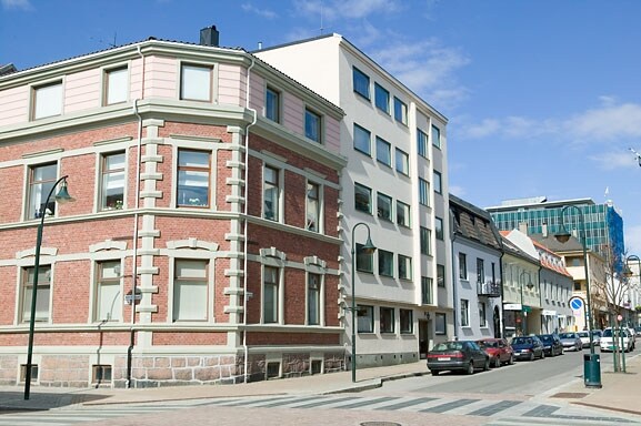 An elegant blend of architecture is displayed, with a historic brick building contrasting against a modern structure. Large windows reflect the clear blue sky, while a wide street is lined with parked cars and lampposts, illustrating the lively atmosphere of Markensgate.