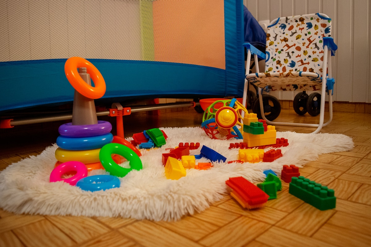 A soft rug on the floor is surrounded by colorful children's toys, including stacking rings, building blocks, and a small rolling toy. A blue playpen and a patterned children's chair are positioned nearby, creating a playful environment suitable for young families.