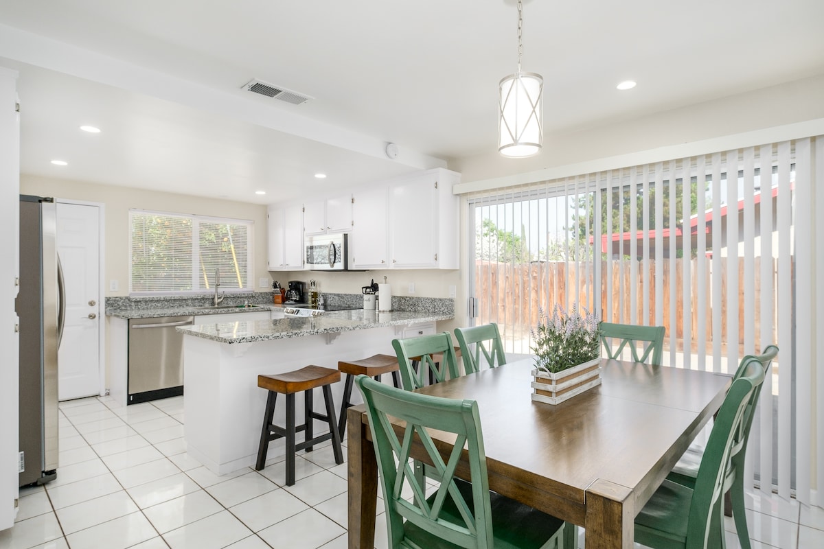 A spacious open kitchen and dining area features a large countertop with modern appliances. A dining table accommodates multiple guests, while natural light enters through large sliding glass doors lined with blinds. The area is finished with white cabinetry and tile flooring.