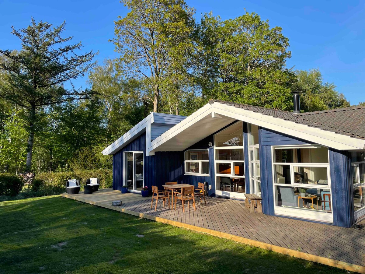 A modern house with a sloped roof features large glass windows that provide ample natural light. Vast wooden deck areas surround the home, with a dining table and chairs situated outside. Lush greenery and trees create a serene atmosphere in the background.