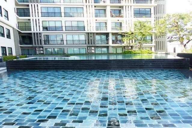 A large outdoor pool is featured with a mosaic tiled design in shades of blue and green. The surrounding area is landscaped with greenery, and several balconies can be seen from the adjacent building, enhancing the tranquil outdoor environment.