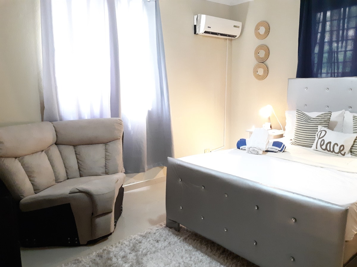 A bright bedroom features a spacious bed with decorative pillows, complemented by a cozy armchair. Natural light filters through sheer curtains, and an air conditioning unit is mounted on the wall. Decorative wall accents add a touch of character.