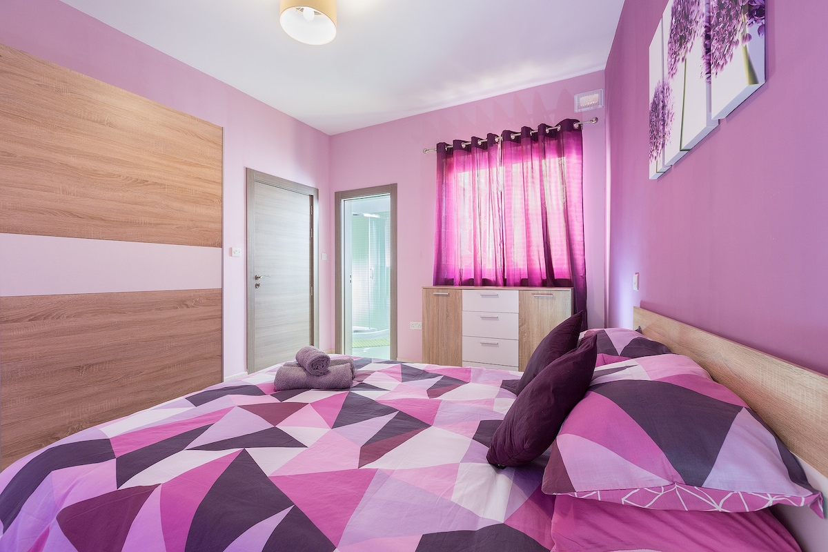 A spacious bedroom features a large bed with a geometric-patterned duvet in shades of pink and purple. Light from the window is filtered through purple curtains. A wooden wardrobe and a dresser are visible, with a towel placed decoratively on the bed.