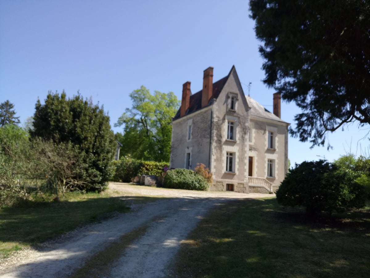 A charming stone house with a classic architectural style is situated at the end of a gravel driveway. Lush greenery surrounds the property, complemented by a variety of trees and shrubs. The clear blue sky provides a serene backdrop to this inviting setting.