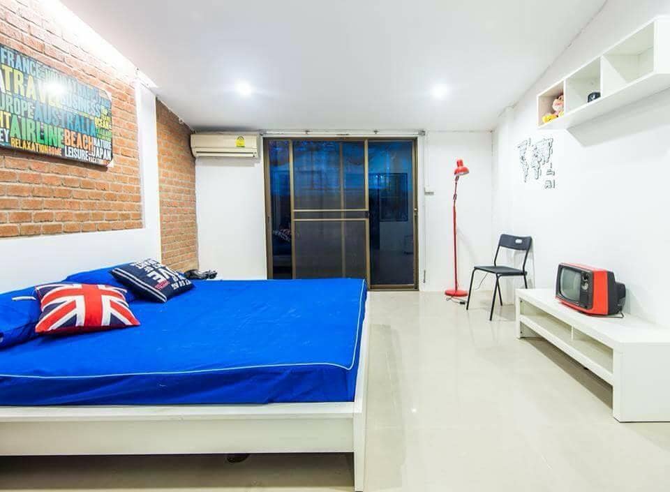 A spacious sleeping area features a large bed with a blue coverlet and decorative pillows. A wooden shelf displays decor items, while a vintage television sits on a white console. Natural light filters through sliding glass doors, complemented by a chair and a reading lamp.