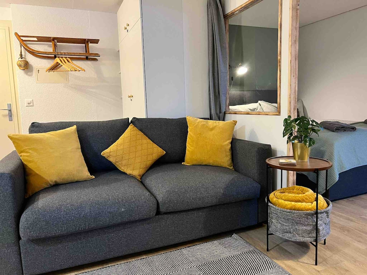 A comfortable seating area is presented with a grey sofa adorned with two yellow cushions. A small side table with a plant is positioned nearby, contributing to the cozy ambiance. In the background, a sleek bed with neutral bedding can be seen, hinting at a versatile living space.