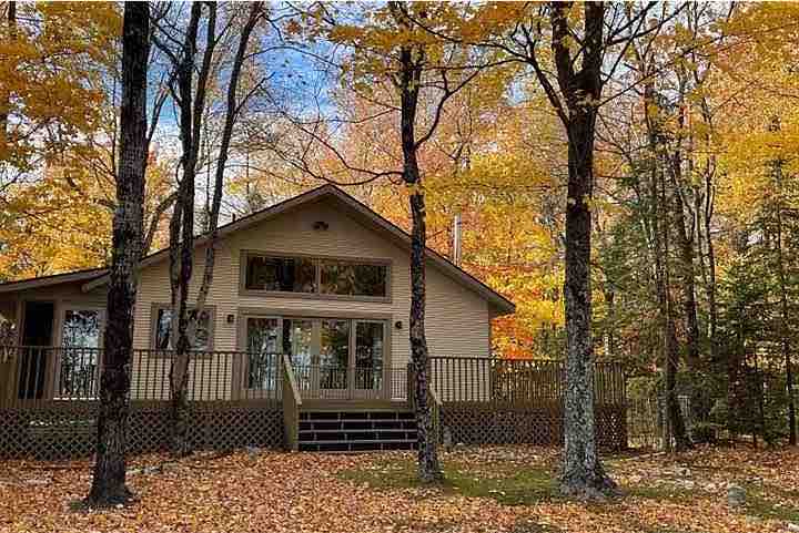 A cozy cottage is surrounded by vibrant autumn foliage, with yellow and orange leaves adorning the ground. A spacious deck extends from the front, featuring large windows that provide a view of the natural landscape. Trees frame the structure, enhancing the tranquil setting.