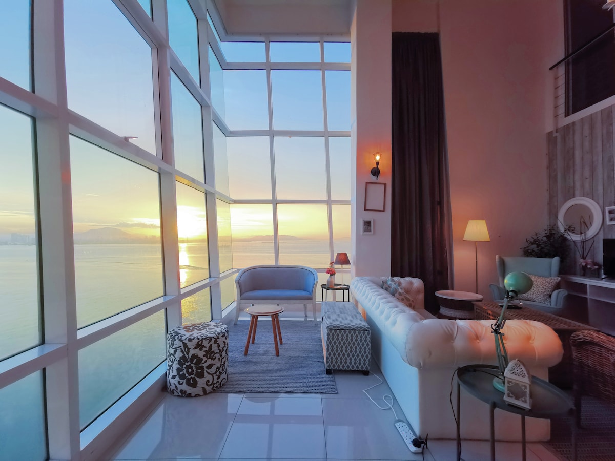 A spacious living area features large windows offering panoramic views of the harbor and sunset. A comfortable sofa is situated beside a small coffee table and a floor lamp, with decorative poufs placed on a soft rug. Natural light fills the room, creating a relaxing ambiance.