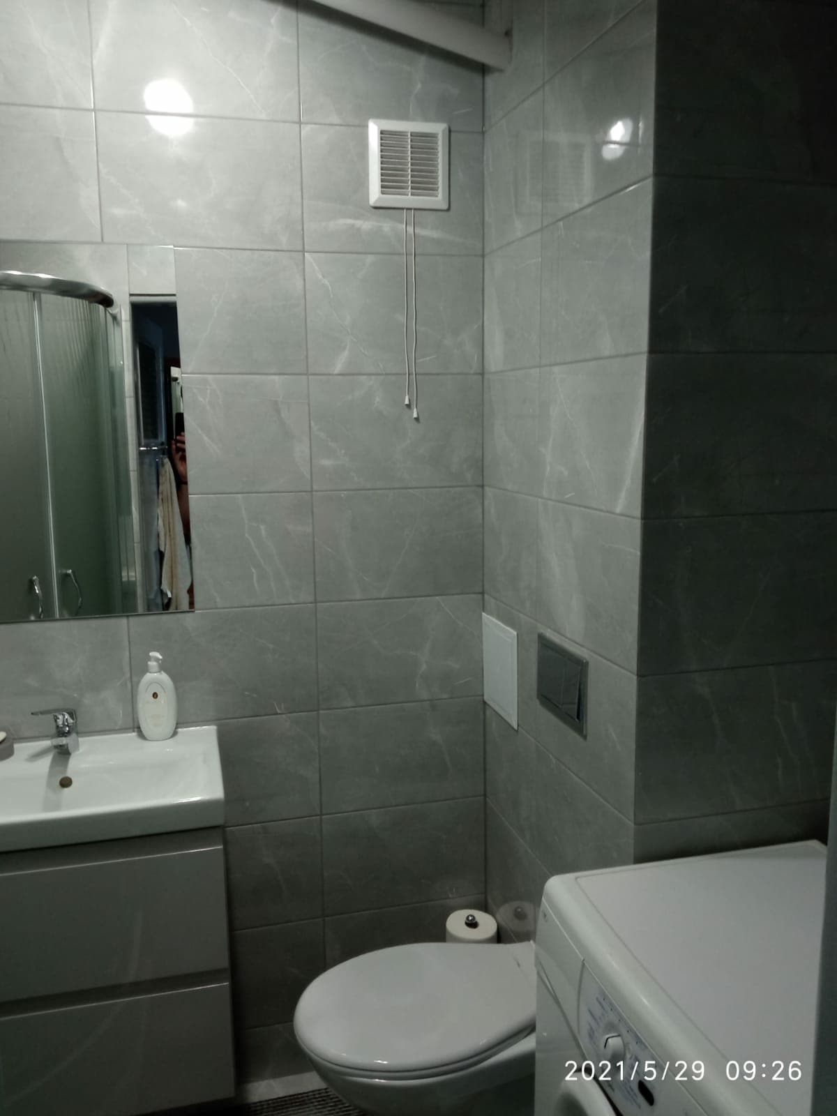 A modern bathroom features grey tiled walls, illuminated by natural light reflecting off the mirror. A compact vanity with a sink and storage is positioned alongside a toilet. A washing machine is located beside the wall, enhancing functionality within the space.