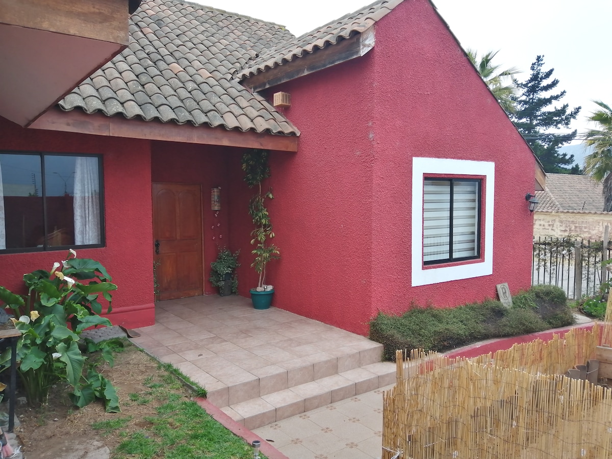The vibrant red exterior of the house contrasts with the tiled roof. A wooden door is framed by a potted plant, while steps lead up to a spacious entrance. Nearby, a garden area features well-kept greenery and a bamboo fence.